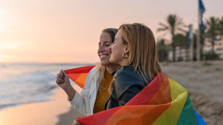 Two women hugging and holding a pride flag on the beach