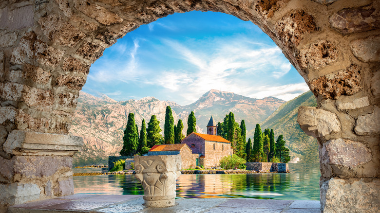 Stone archway with fountain framing a view of mountains and red roof building surrounded by tall trees