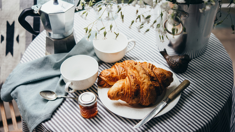 A striped café table with croissants, a french press, coffee cups, and flowers