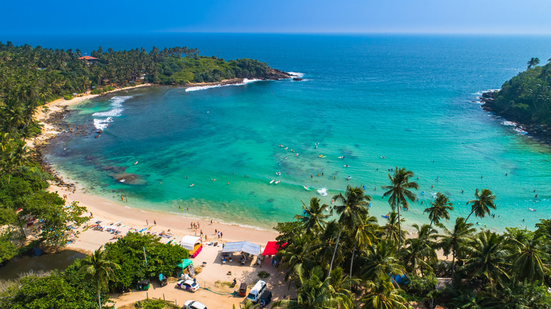 An aerial view of Hiriketiya beach in Sri Lanka with huts and sun lounges on the sand and surfers in the sea