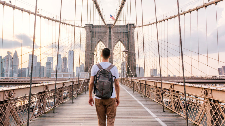 Man with a backpack crosses a large bridge to New York City with an American flag in the distance