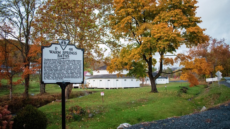 A white and black sign to Warm Springs Pools with fall trees and a bath house in the background