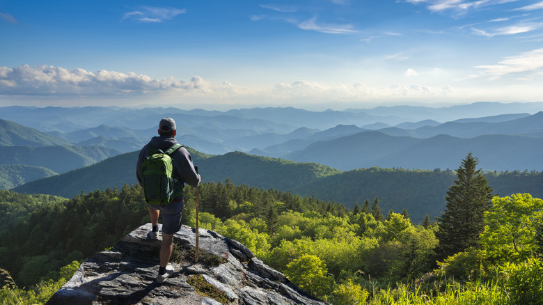 A man stands on a ledge looking out over endless green hills and mountains in the misty distance