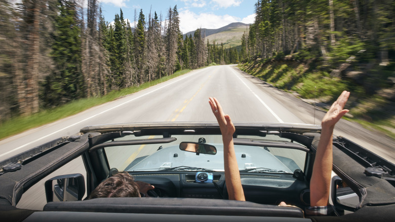 Two people driving in a car down a paved road with trees blurring on the sides and mountains in the distance