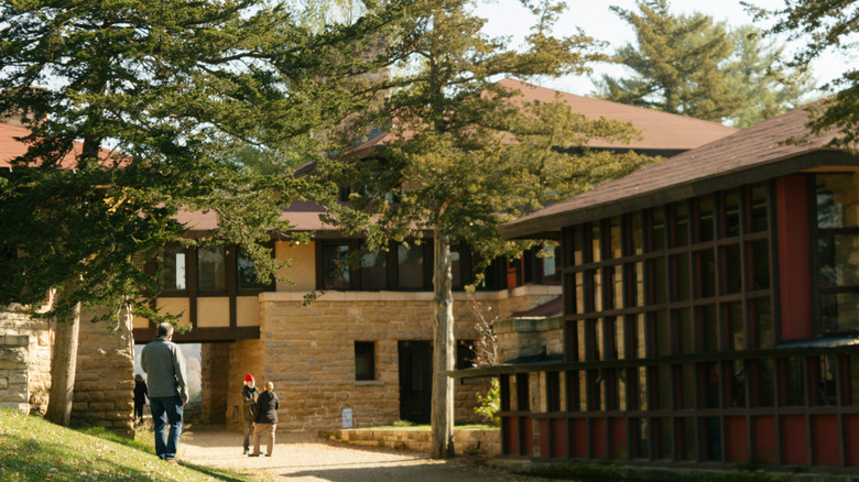 A small group of people walk in front of a stone and wood building with trees in its garden