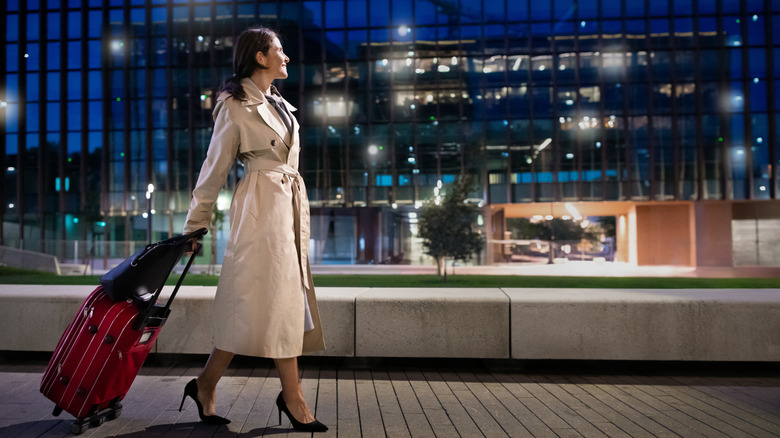 A woman in a trench coat wheeling a red suitcase in front of a building at night