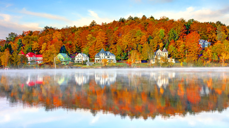 View from the water of Saranac Lake with houses lining the water, and autumn trees with hues of orange, green, and red behind the houses.