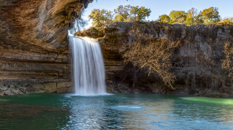 A waterfall in Dripping Springs, Texas pours into a pool over sand-colored stones with trees on a ridge in the background