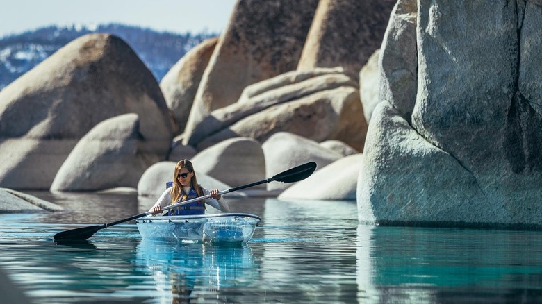 a woman paddles a clear kayak in front of giant boulders