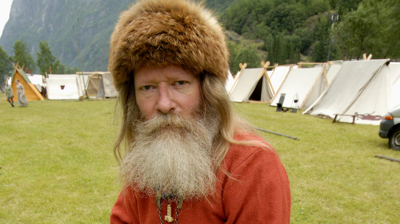 A man with a long pale beard and fur hat stands in front of traditional Viking tents.