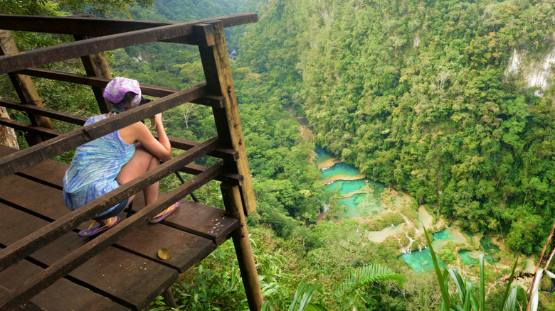 a woman knealing down on a wooden platform to take a picture of Semuc Champey from above