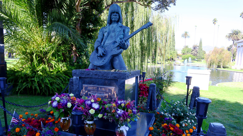 Grave with statue of Johnny Ramone with flowers surrounding it in a peaceful park with pond