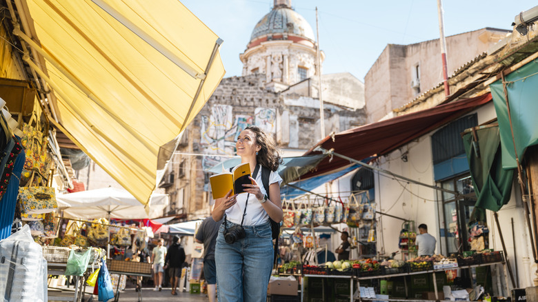 A solo traveler walking through a market holding a guidebook