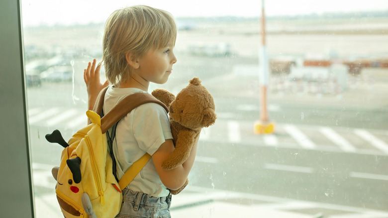 A small child holding a teddy bear while staring at the tarmac out a window