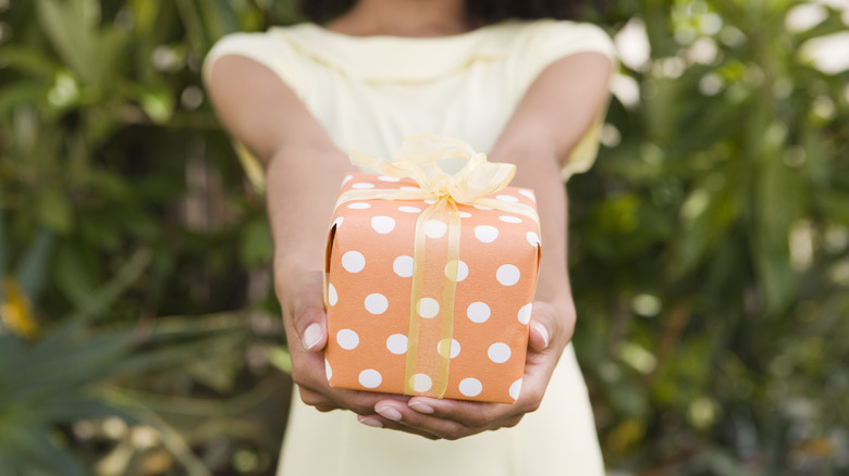 A woman holding out a wrapped gift while stood outside.