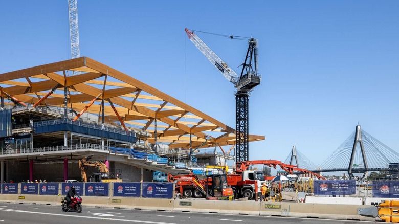 Construction site featuring cranes, barriers, trucks, and an angular, wooden roof skeleton