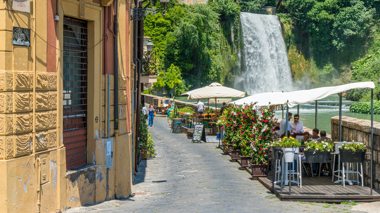 outdoor cafes line a small street on a river bank with a large waterfall in the background