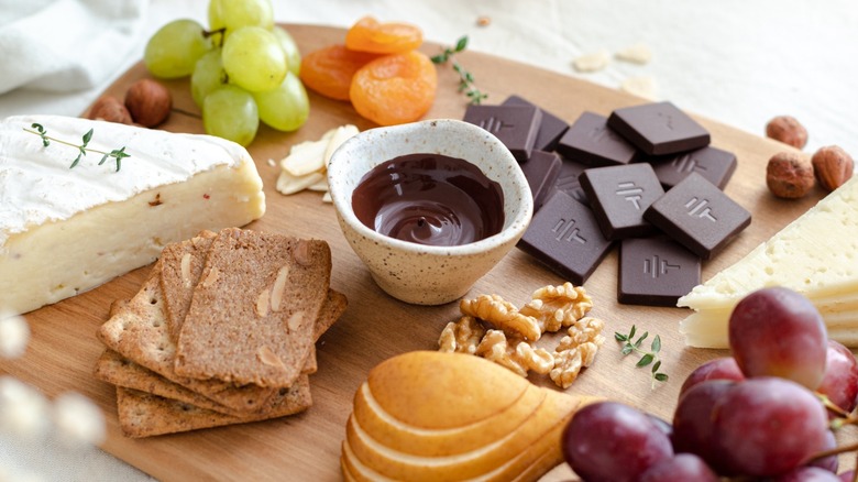 A charcuterie board filled with To'ak chocolate pieces, crackers, cheese, grapes, apricots, and other food