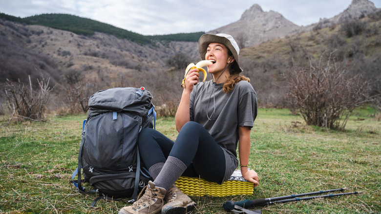 A female hiker sitting on a hilltop with gear, eating a banana.