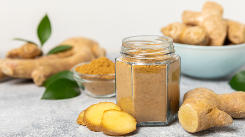 Ginger powder in a jar surrounded by cut pieces and whole ginger roots.