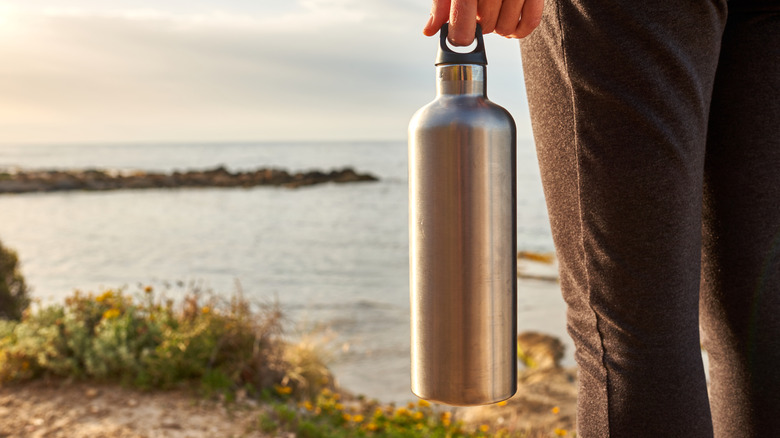 A hiker's hand holding an aluminum bottle of water standing on a hill.