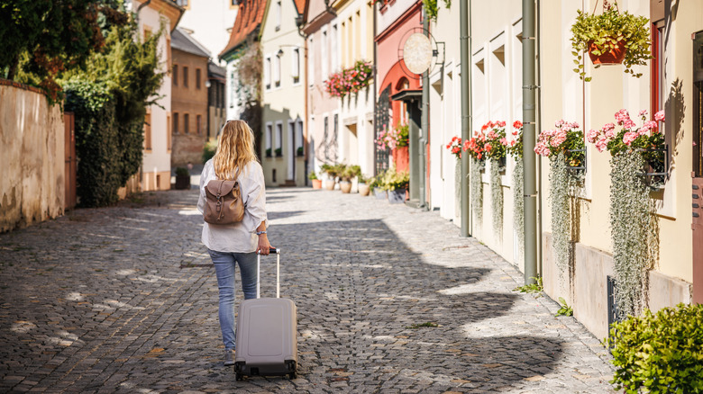 Single woman pulling suitcase along cobbled stone street lined with flowers