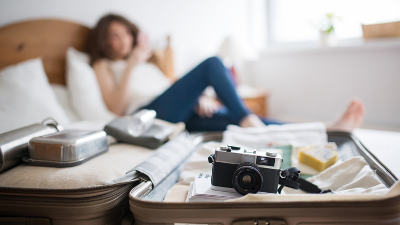 Unpacked suitcase with camera and single woman looking at phone in bed