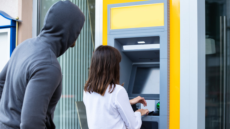 A thief behind a woman withdrawing money from an ATM