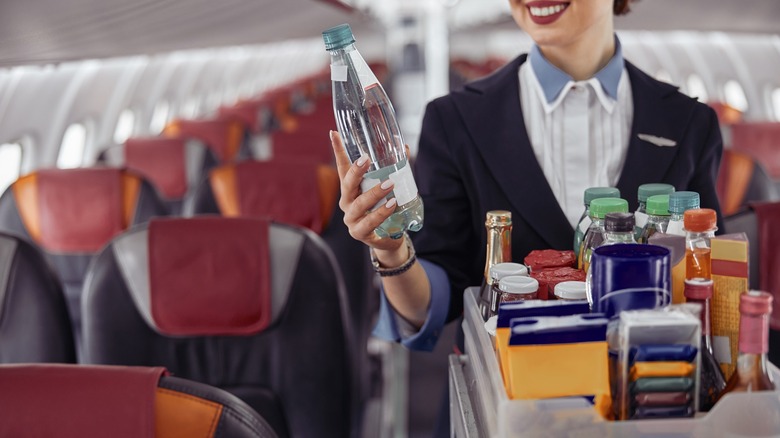 flight attendant holding water bottle standing behind drink trolley in airplane