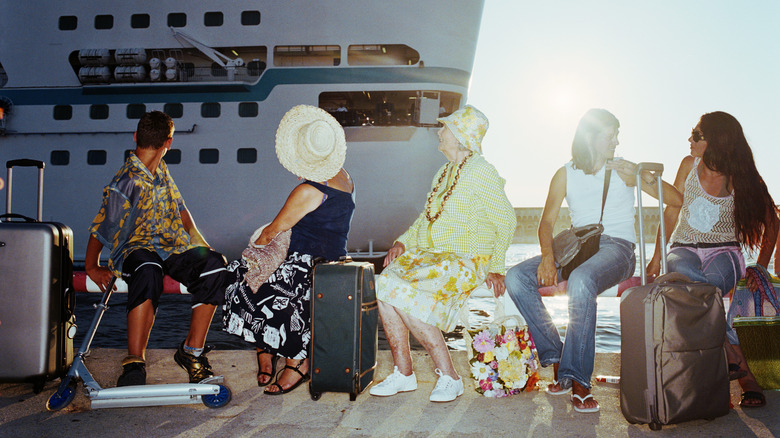 Passengers sitting on rolling suitcases, staring at a cruise ship behind them in port