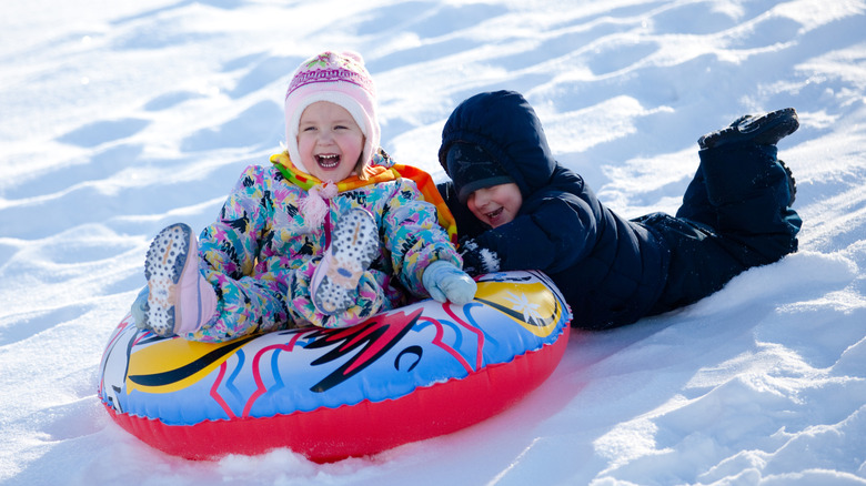 Two kids sledding down a snowy hill, one in a tube and one holding on