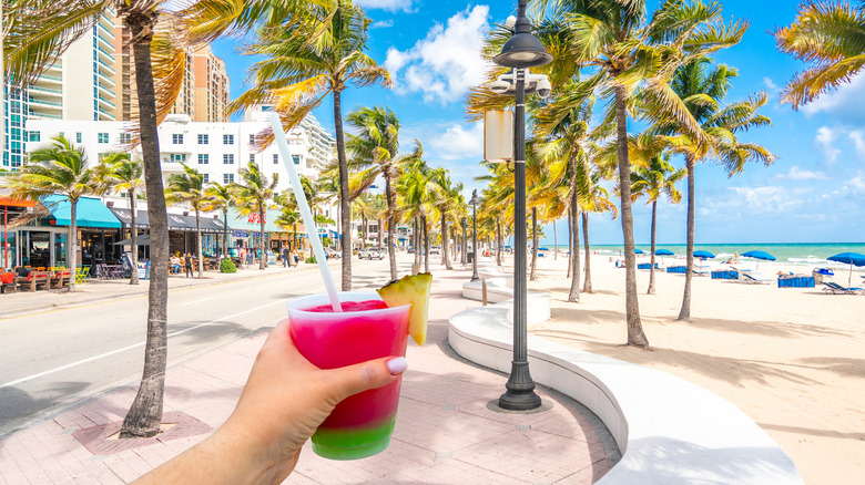 A pink and green drink being held in front of a Fort Lauderdale beach, featuring palm trees, shops, and blue umbrellas