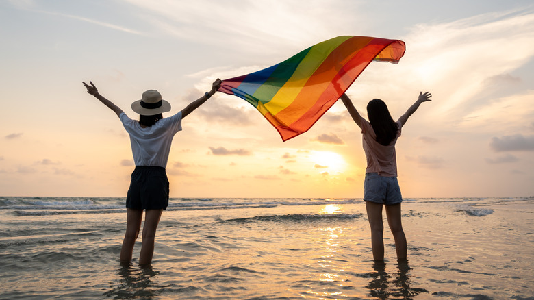 Two women holding a pride flag with their arms raised on the beach