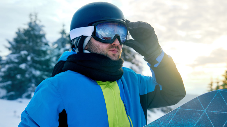 Man with full gear standing on top of a mountain with a snowboard, staring at the sight before him