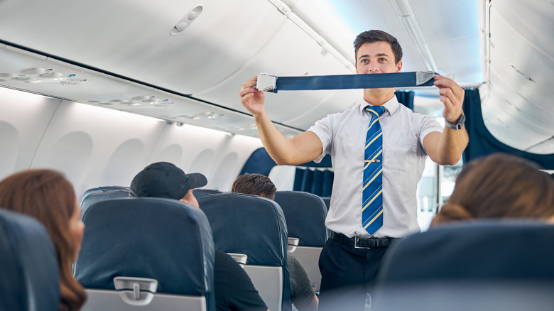 A flight attendant with bare arms and no tattoos shows how to buckle seatbelt