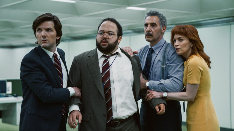 Four characters in an office building huddle together under stress and concern