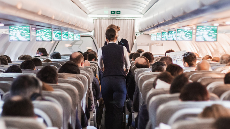 Interior of aircraft mid-flight with passengers seated and cabin crew walking through the aisle