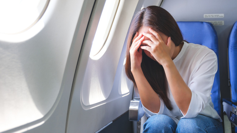 Woman sitting in window seat on plane holding her head in her hands