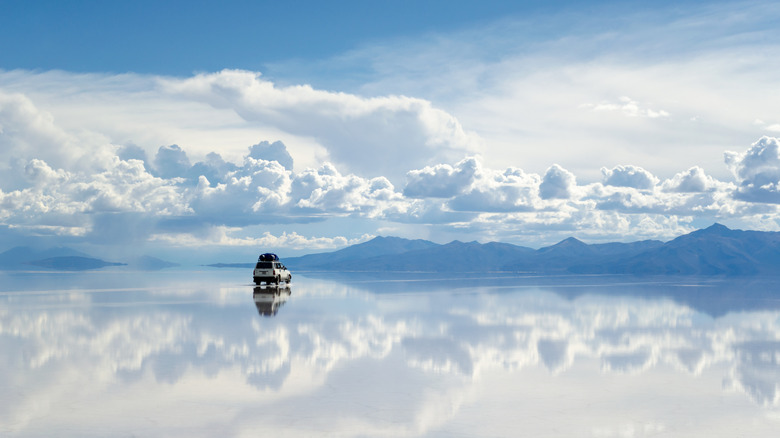 A white car sits on the reflective surface of a shallow lake with mountains and clouds in the distance and sky above