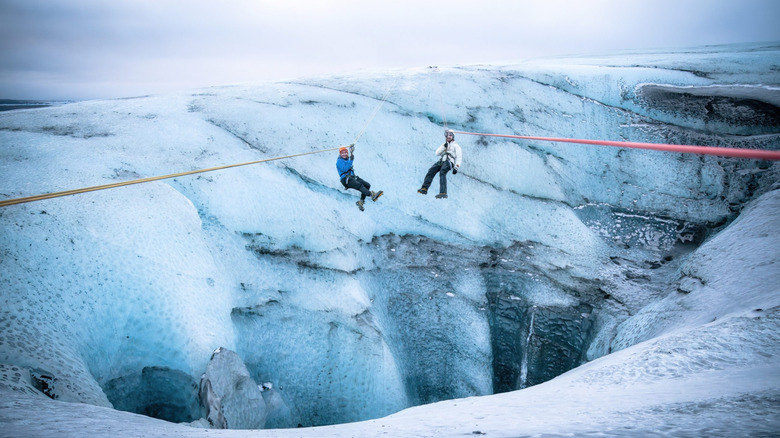 two men hang on separate zip lines over the open crevasse of an icelandic glacier