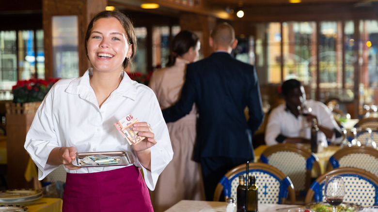 Server in restaurant smiling holding tip tray and euro notes as couple walk behind her