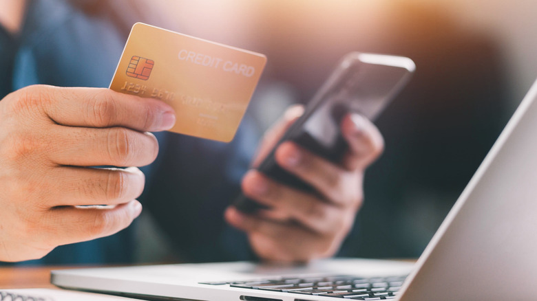 A person holding a gold credit card and a phone in their left hand a few inches in front of an open laptop screen