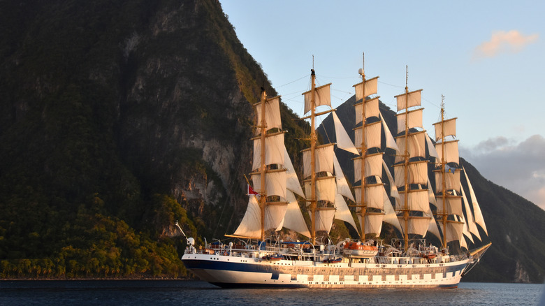 A five-mast tall ship in water sailing past a mountain