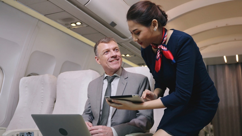 Woman flight attendant in blue dress with tablet in hand helps seated man on plane