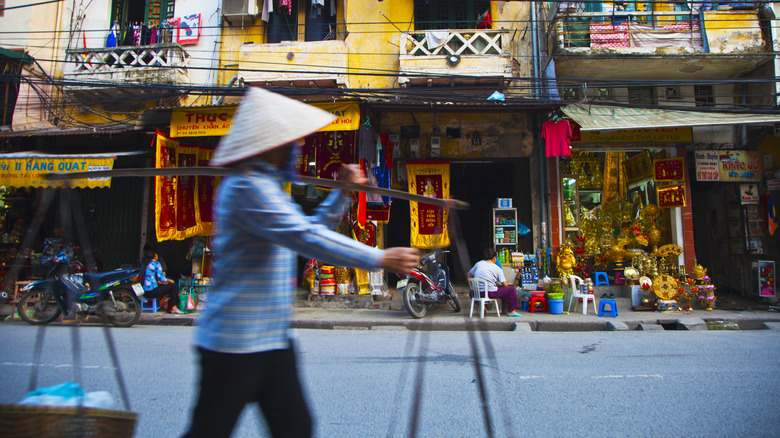 Street vendor in straw hat walks by traditional shop houses in Hanoi